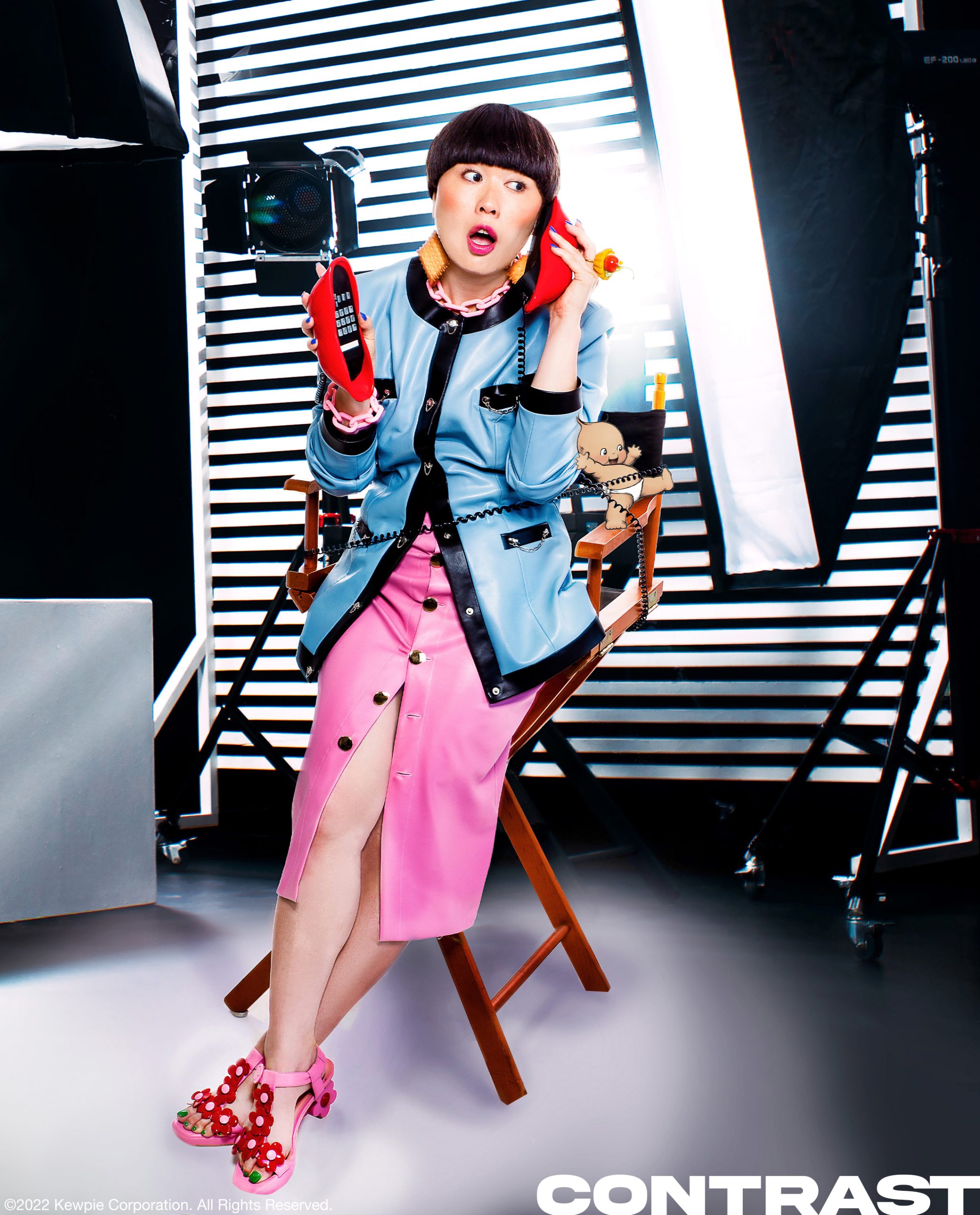 Comedian and HBO MAx star Atsuko Okastuko poses in a director's chair wearing blue blazaer jacket and a pink skirt holding a red lips house phone and surrounded by stage production lights. The image features an illustrated dark tone Kewpie doll character. This editorial was produced by ONCH, formerly known as Onch Movement.