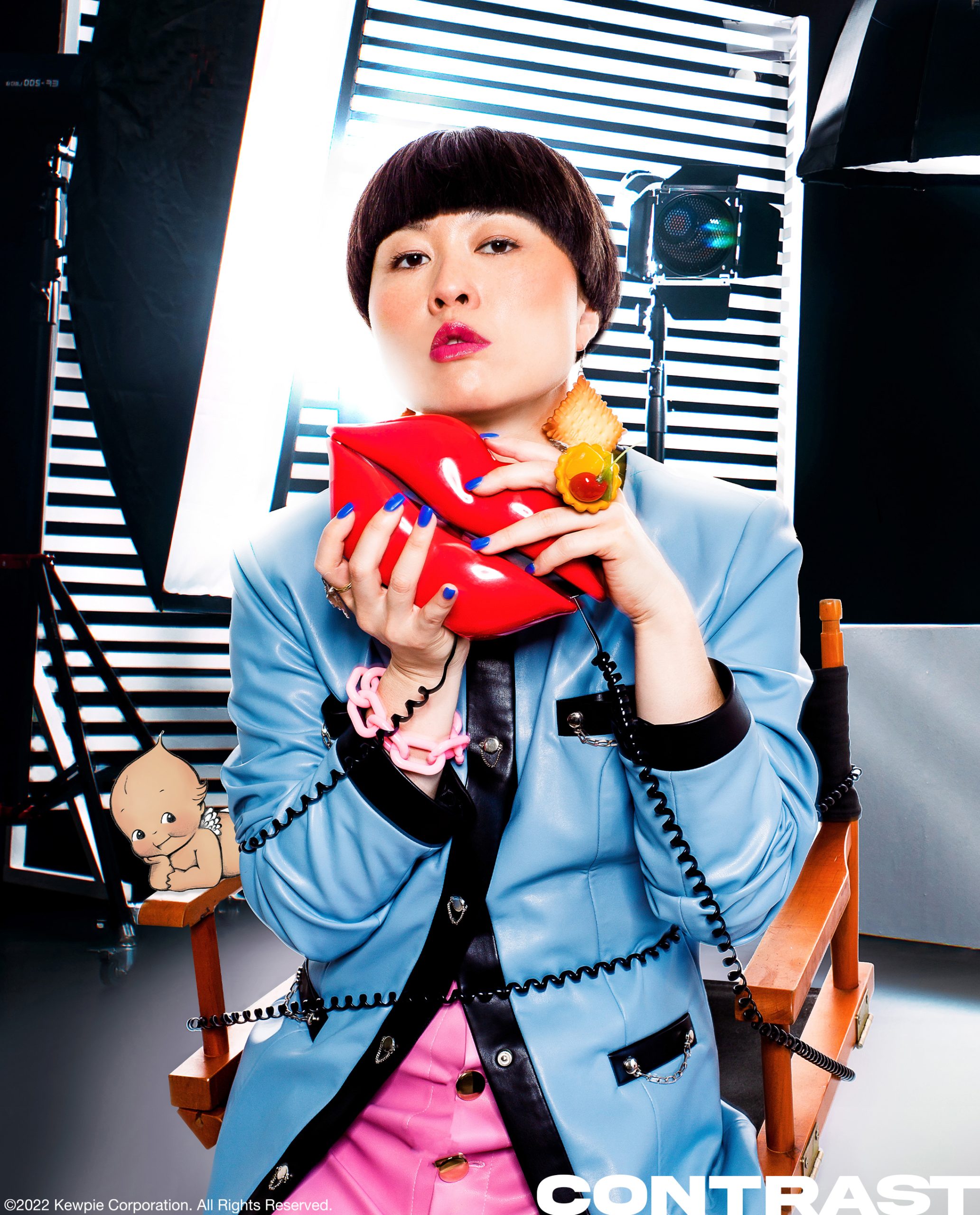 Comedian and HBO MAx star Atsuko Okastuko poses in a director's chair wearing blue blazaer jacket and a pink skirt holding a red lips house phone and surrounded by stage production lights. The image features an illustrated dark tone Kewpie doll character. This editorial was produced by ONCH, formerly known as Onch Movement.