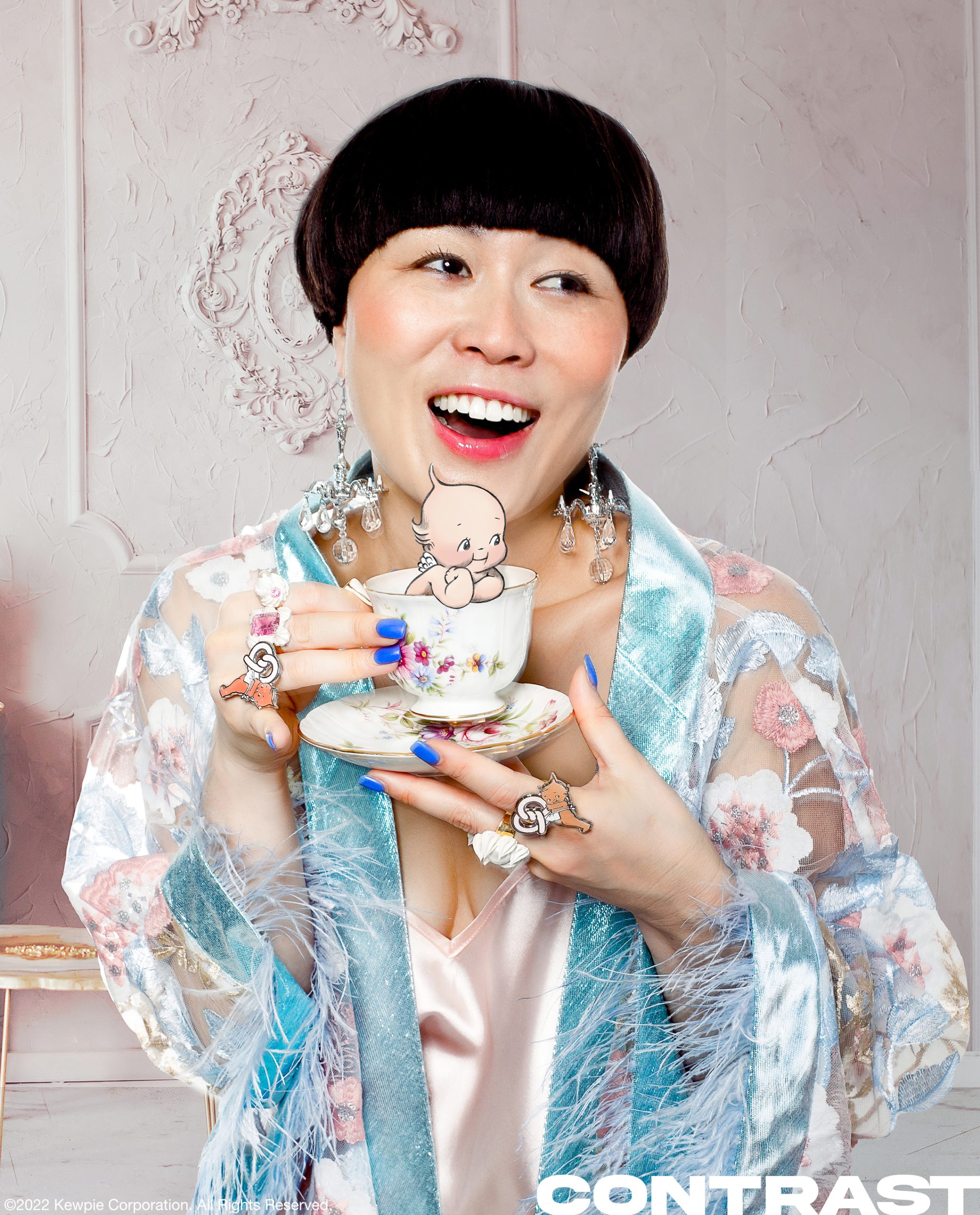 Comedian and HBO Max star Atsuko Okastuko poses in a floral blue silk robe and a pink silk slip holding a floral tea cup while laughing and smiling and wearing chandelier ear rings. The image features an illustrated light tone Kewpie doll character. This editorial was produced by ONCH, formerly known as Onch Movement.