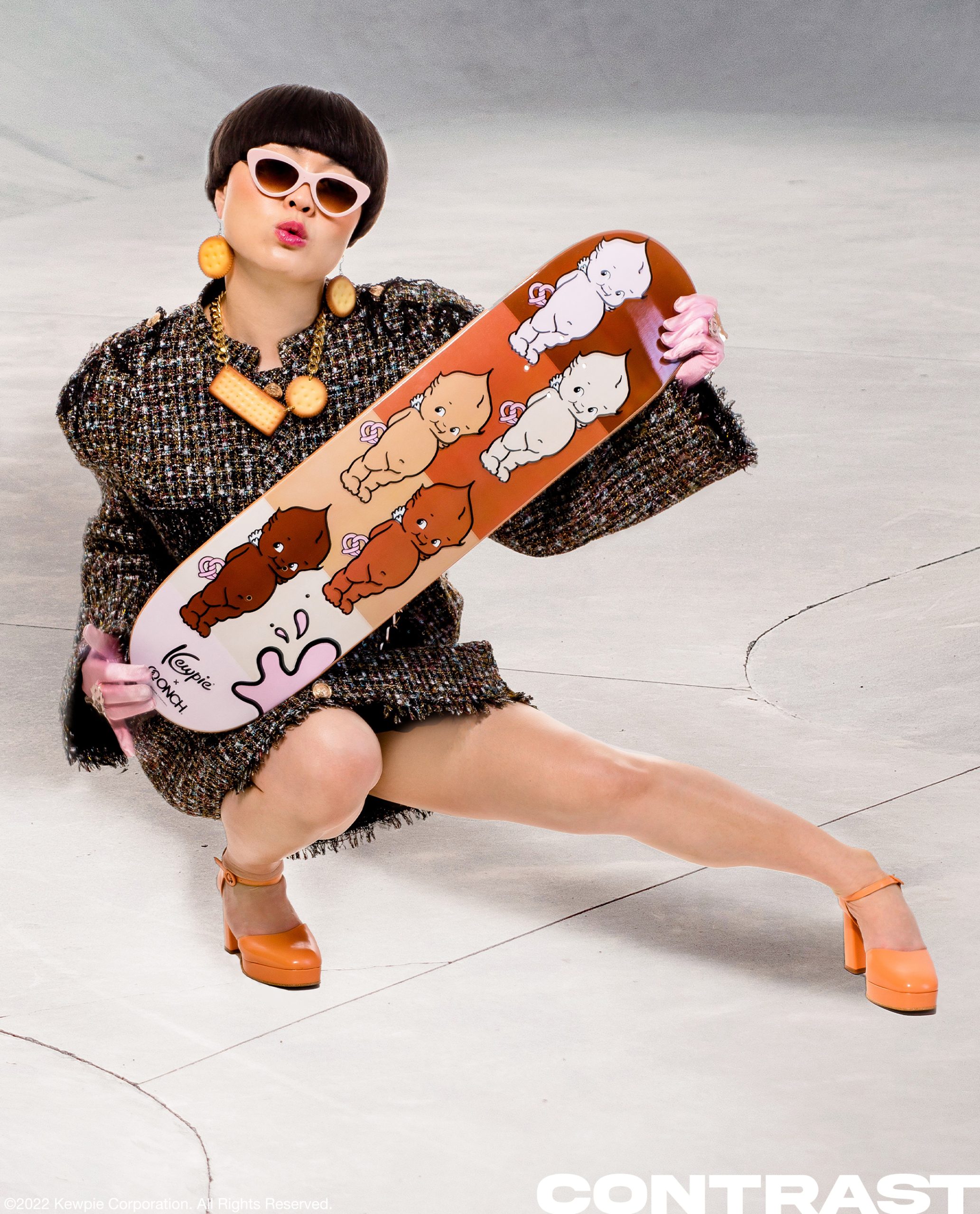 Comedian and HBO Max star Atsuko Okastuko poses in a black jacket and orange retro strap on heels while holding an ONCH and Kewpie skateboard deck in the middle of a skateboard park. The image features an illustrated light tone Kewpie doll character. This editorial was produced by ONCH, formerly known as Onch Movement. Atsuko is also wearing a cracker/cookie necklace designed by ONCH.