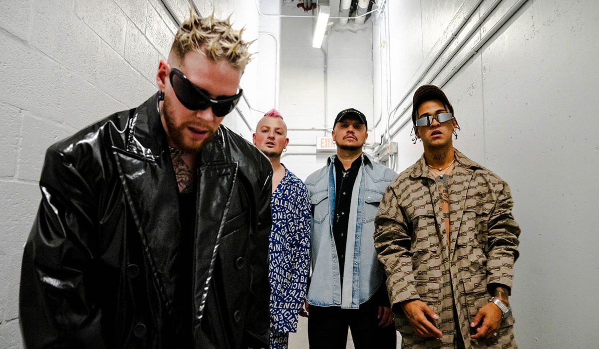 All four members of the Colombian boy band Piso 21 poses in a hall way. They are each wearing a stylish jacket in black leather, blue Balenciaga print, blue denim and brown.